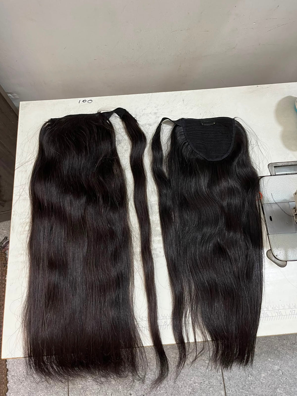 Two black hair extensions laid flat on a white surface, one with a clip-in style attachment and one with an open lace top, shown against a plain indoor background.