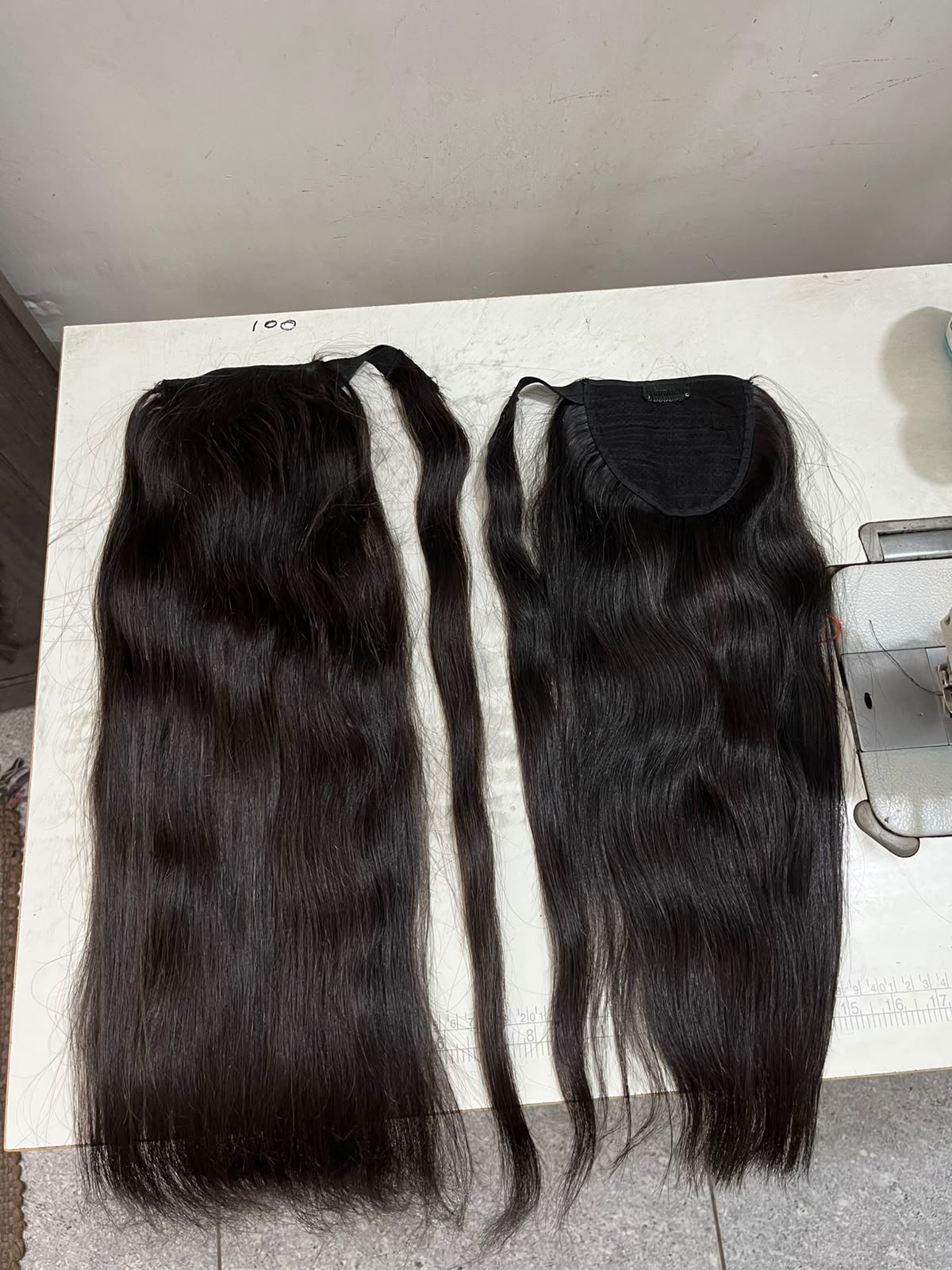 Two black hair extensions laid flat on a white surface, one with a clip-in style attachment and one with an open lace top, shown against a plain indoor background.